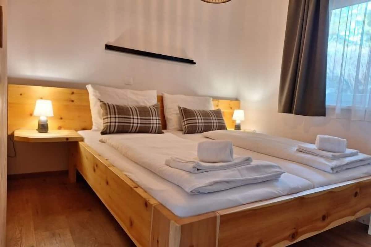 A double bed with white linens and two pillows, a wooden headboard, bedside lamps, and a window with curtains.