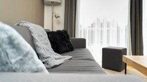 A modern living room with a grey sofa, a black throw pillow, a grey blanket, a floor lamp, and a view of a cityscape through a large window.