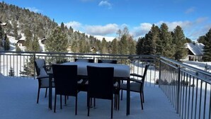 A snow-covered outdoor dining area with a table and chairs, surrounded by a metal railing, overlooking a forested mountain.