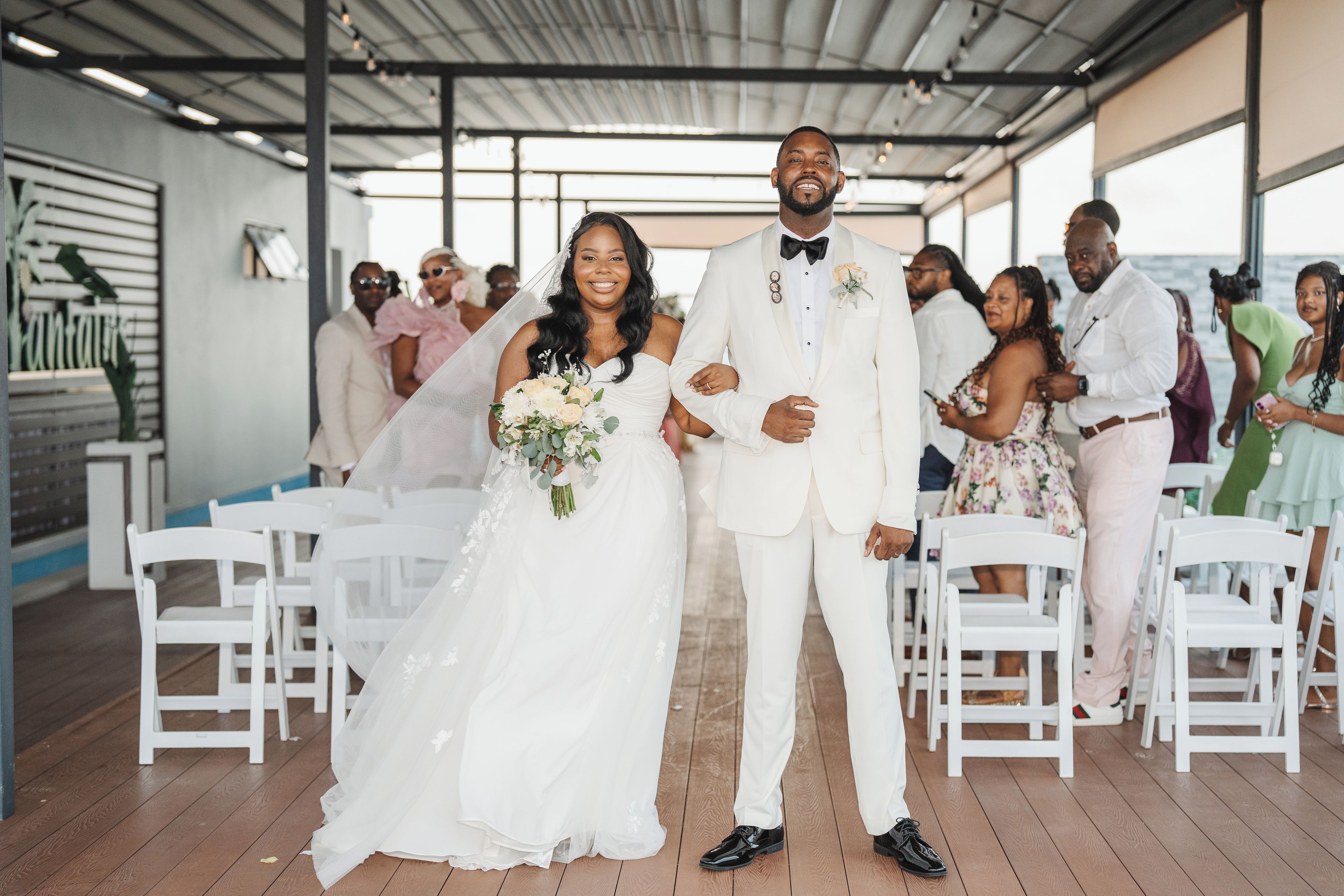 A wedding ceremony with a couple standing at the aisle's center, surrounded by guests seated on white chairs.