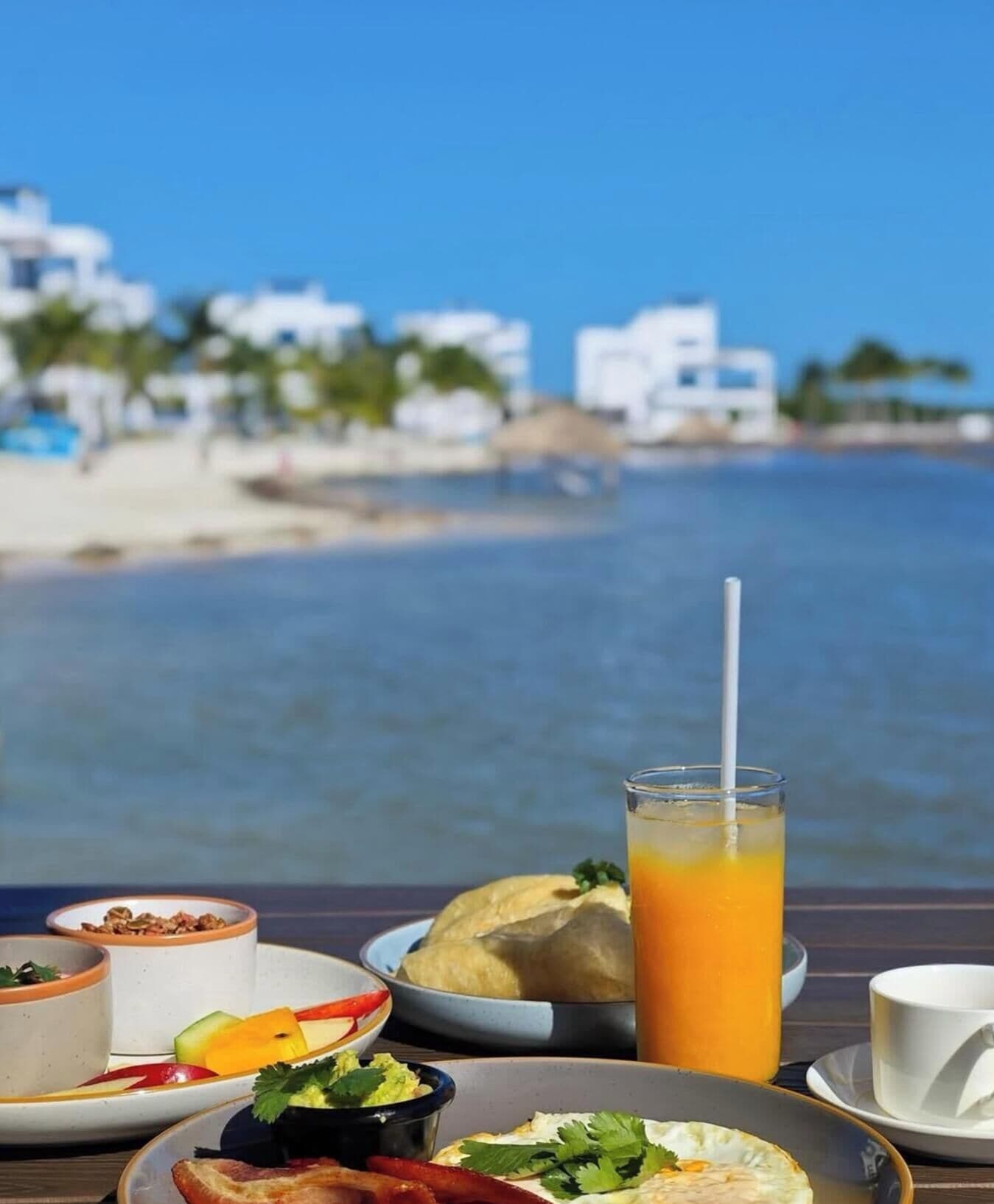 Una tavola imbandita con prodotti per la colazione, tra cui uova, pane tostato e un bicchiere di succo d'arancia, con vista su una spiaggia e edifici bianchi con palme.