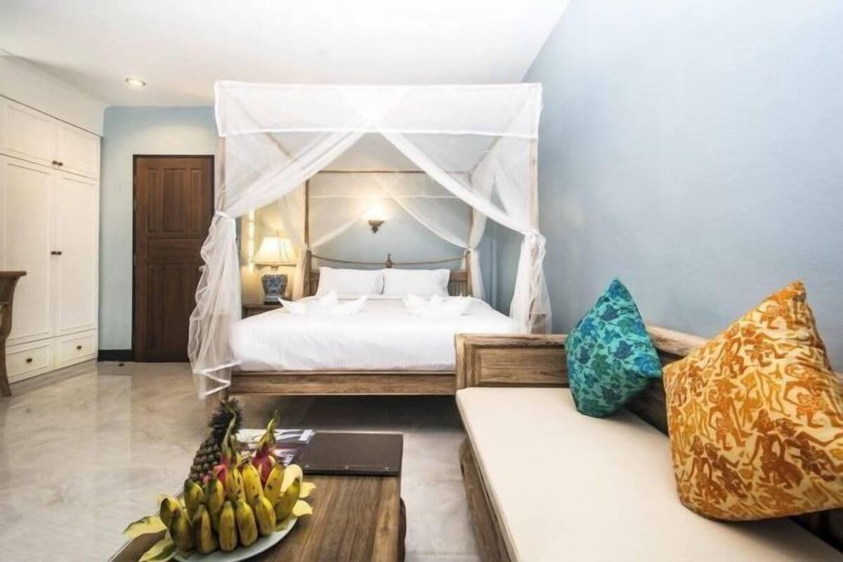 A bedroom with a canopy bed, a bedside table with a fruit bowl, and a sofa with patterned cushions.