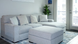Un sofá seccional gris con almohadones a cuadros, un reposapiés a juego, un tapete con estampado azul y blanco, una planta en maceta y vista a un paisaje urbano a través de una puerta con persianas.