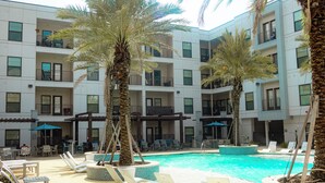 Un área de piscina con sillones de descanso y palmeras, rodeada por un edificio de varios pisos con balcones.