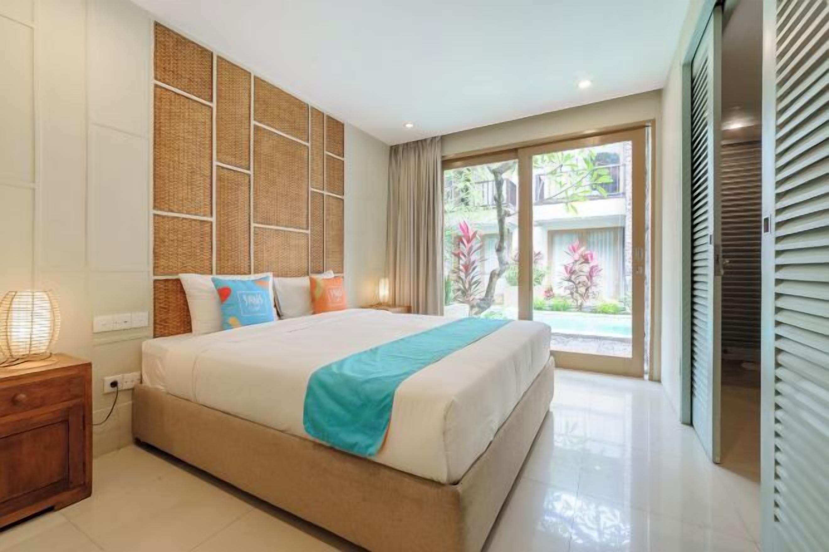 Een hotelkamer met een groot bed, een nachtkastje, een wand met geweven paneeldecoratie en uitzicht op groen door een schuifdeur.