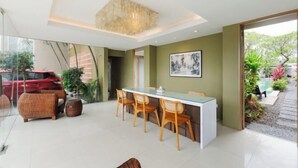 A modern dining area with a glass table and wooden chairs, a large painting on the wall, and a view of the outdoors. - Sans Vibes Astana Residence Seminyak (Bali)