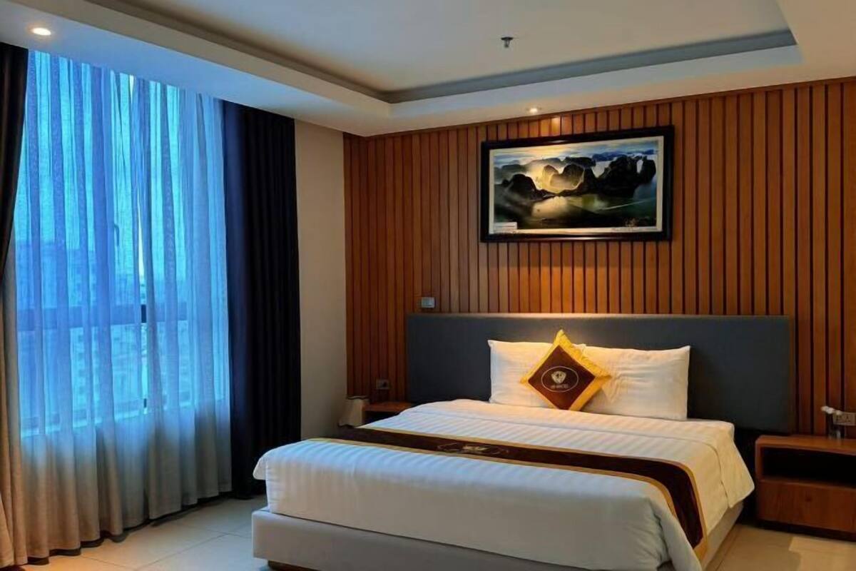 A hotel room with a large bed, a headboard with a painting, a nightstand, and a window with curtains.