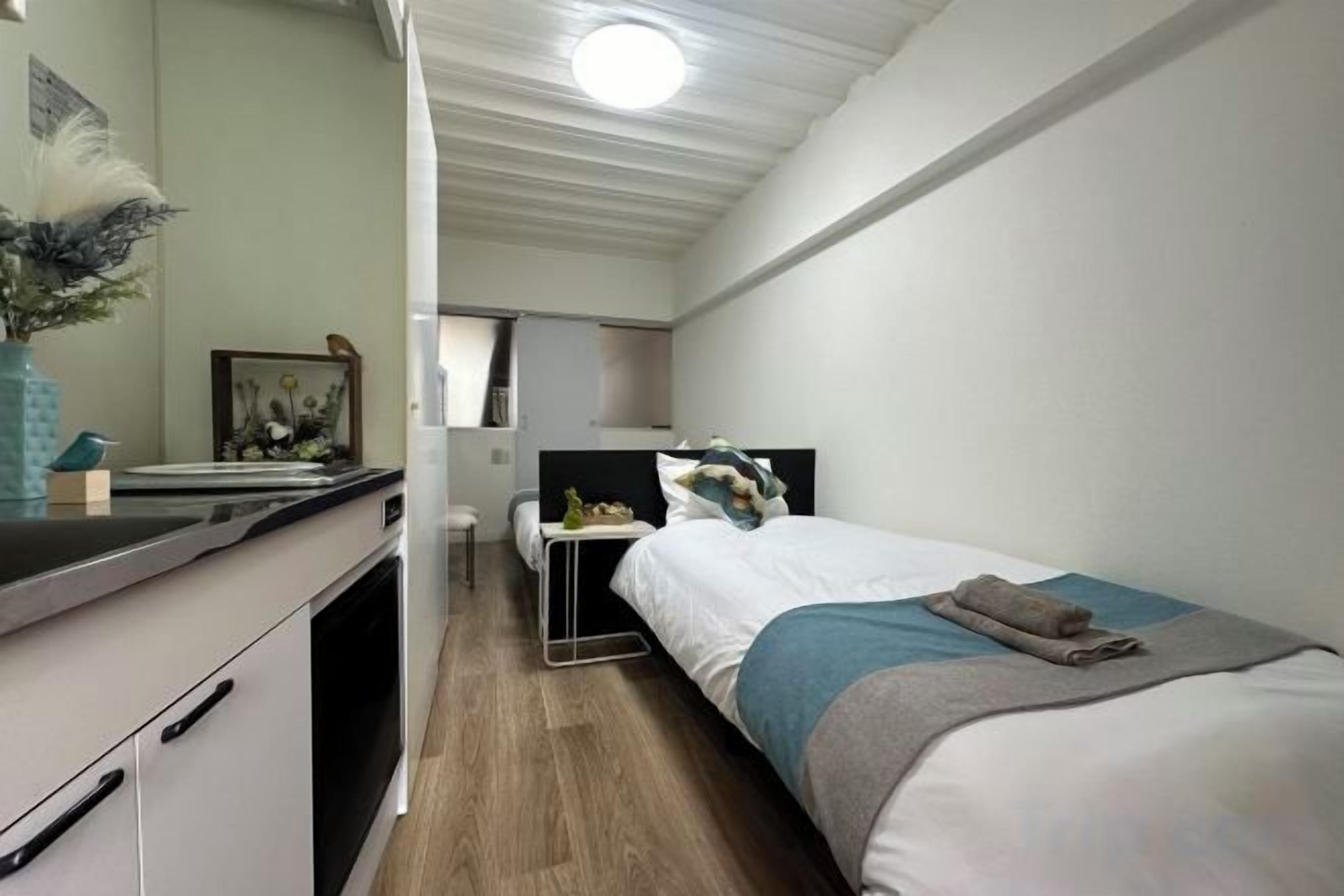 A compact bedroom with a single bed, a bedside table, a mirror, and a small desk with a chair.