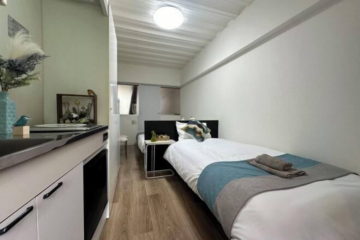 A compact hotel room with a kitchenette, a bed with a blue and grey bedspread, a small bedside table, and a mirror on the wall.