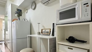 A compact kitchen with a white refrigerator, a microwave, a small table with a 'welcome' sign, and a pair of headphones on a shelf. - 3 Guests Kitchen & Washer 7-min Walk to Waseda University (Tokyo)