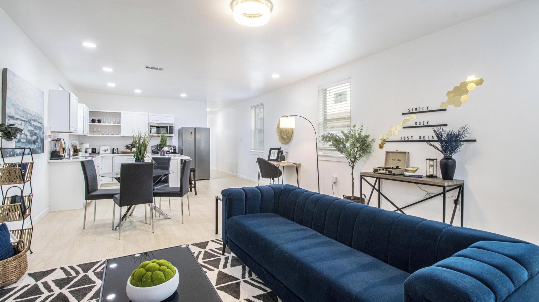 Una sala de estar moderna con un sofá azul, un comedor con mesa y sillas, y una cocina con armarios blancos.