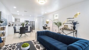 A modern living room with a blue sofa, a dining area with a table and chairs, and a kitchen with white cabinets.