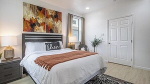 A neatly arranged bedroom with a bed, two bedside tables, a window with curtains, a door, and a wall-mounted artwork.