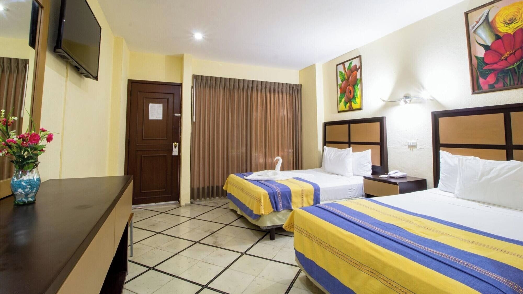 A hotel room with two beds, a TV, a desk with a vase of flowers, and a painting on the wall.