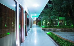 Corredor de hotel com portas de vidro, piso ladrilhado e palmeiras iluminadas por luz verde.