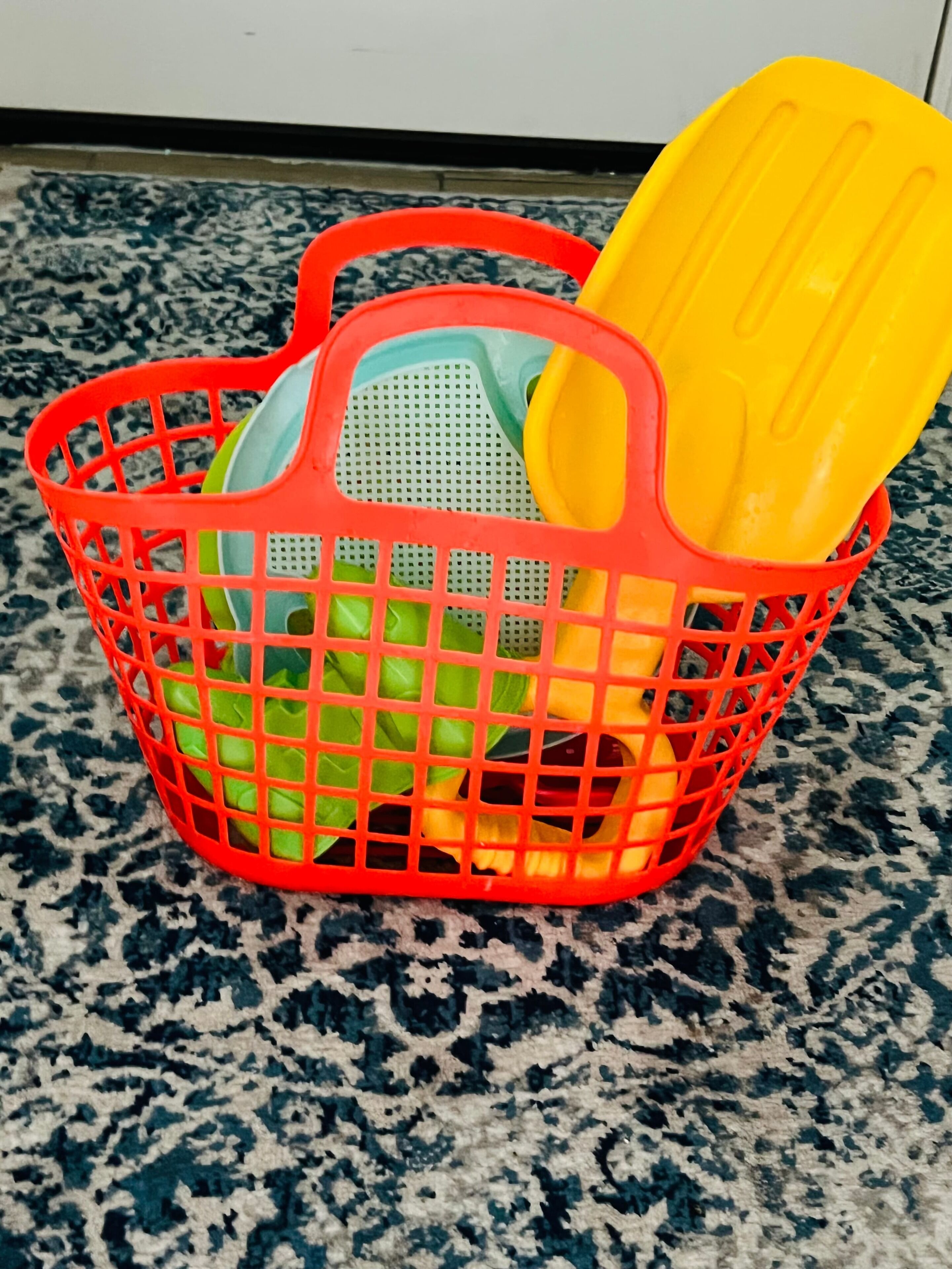 A red plastic basket with a green and yellow lid, placed on a patterned carpet.