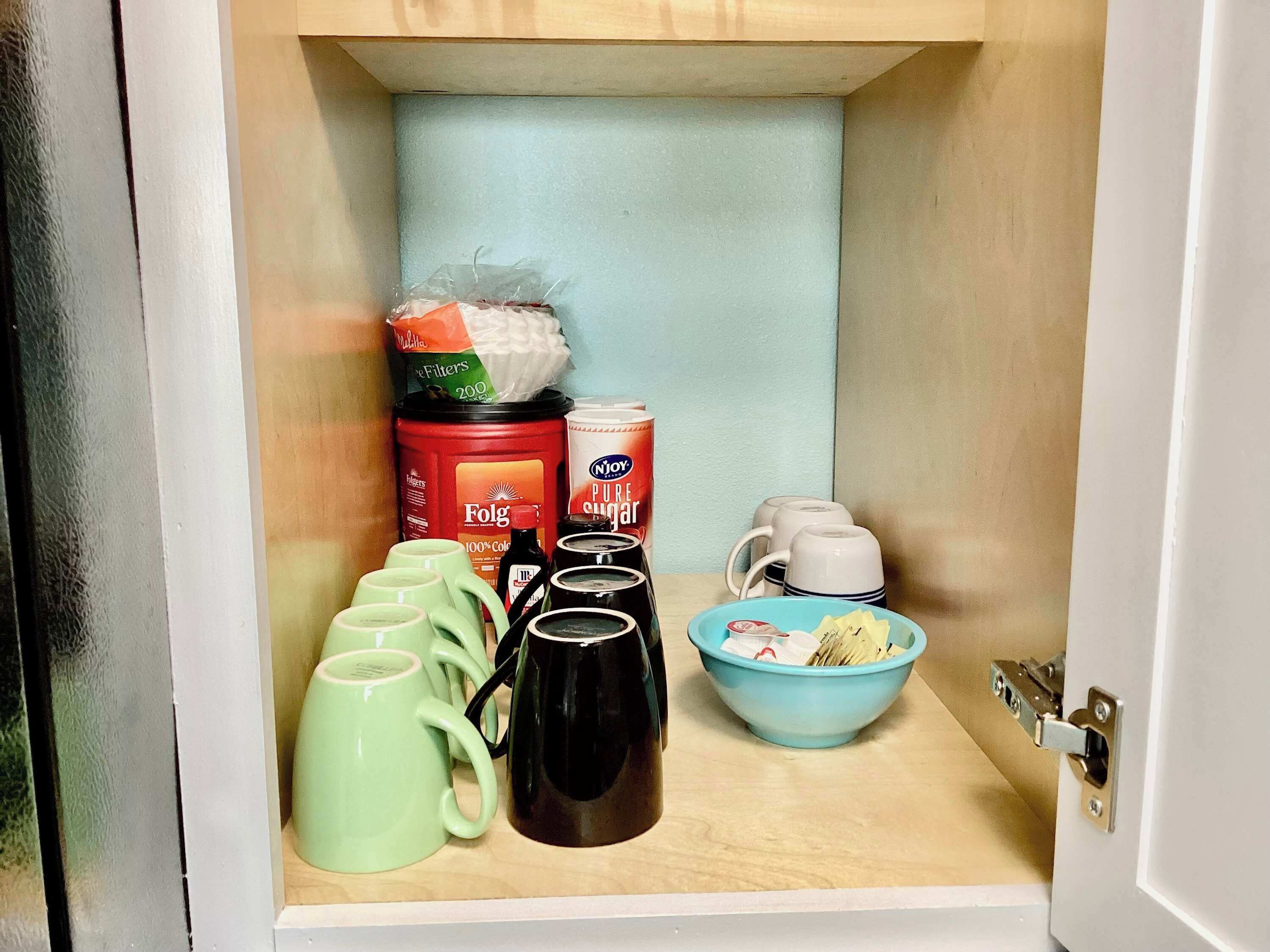 Een keukenkastje met groene mokken, een zwarte koffiepot, een blauwe kom en diverse keukenbenodigdheden.