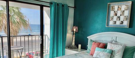 A bedroom with a bed, a nightstand, a lamp, and a view of the beach.