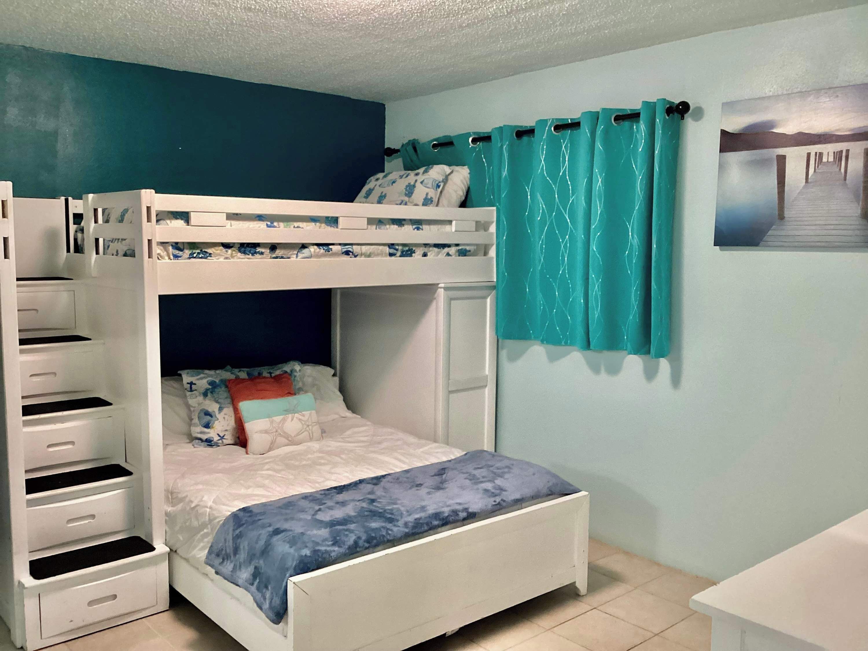 A bunk bed with a lower bunk, a white dresser with drawers, a bed with a blue blanket, and a teal curtain.