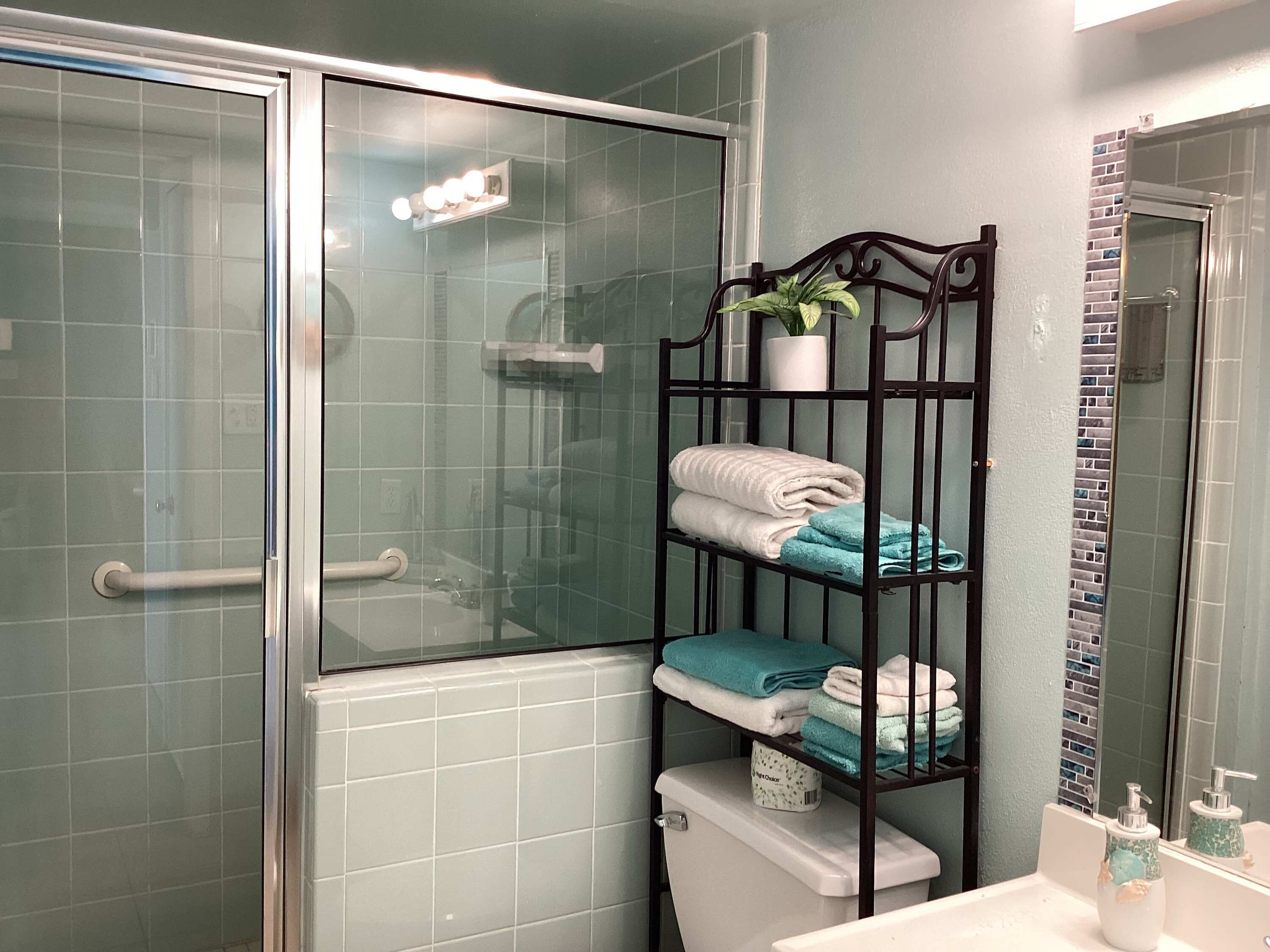 A bathroom with a glass shower enclosure, a toilet, a sink with a mirror, and a shelf with folded towels.
