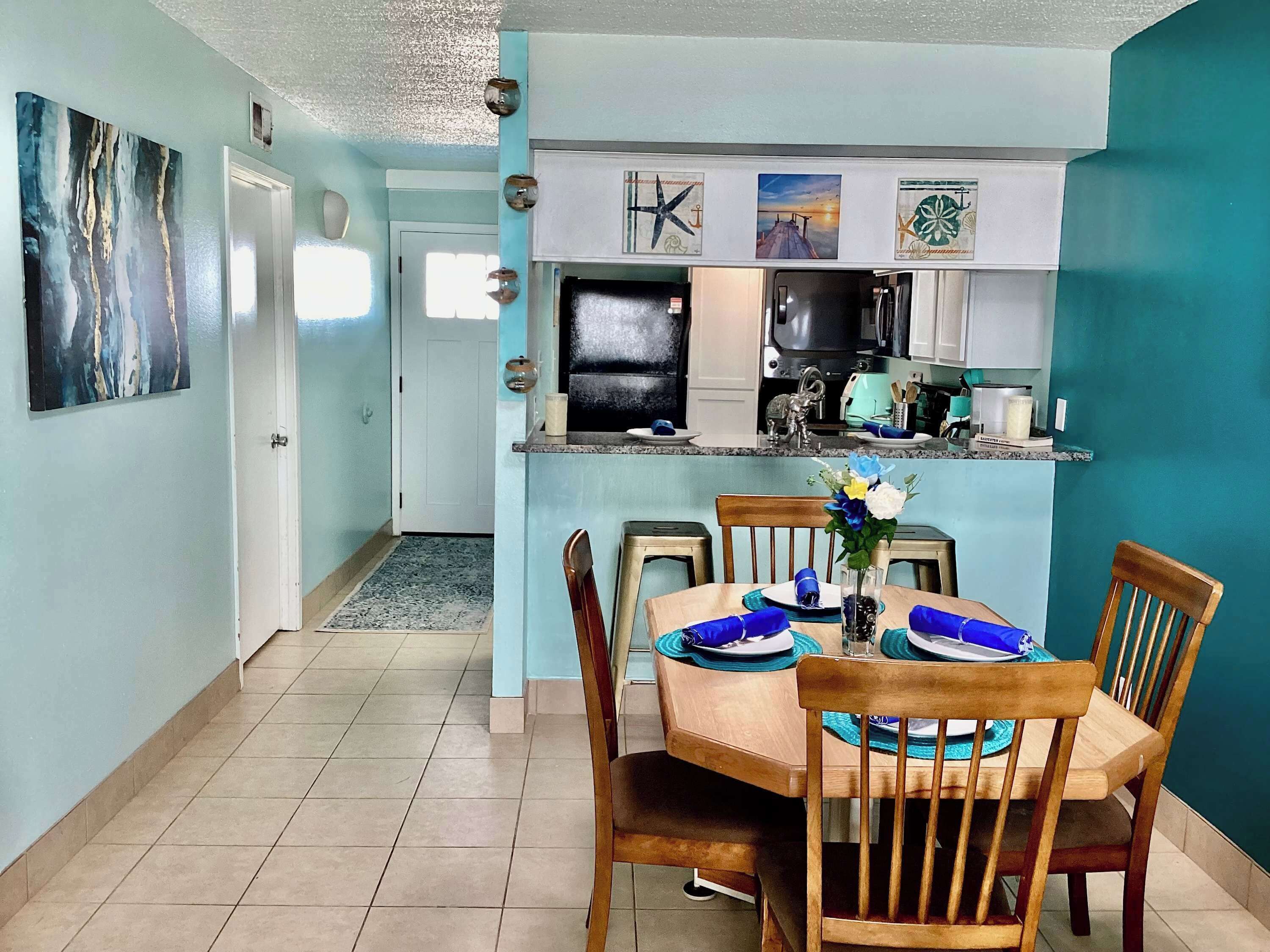 A dining area with a table set for four, a kitchen in the background, and a hallway leading to a door.