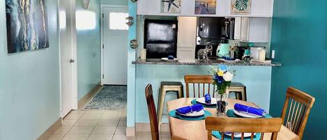 A dining area with a table set for four, a kitchen in the background, and a hallway leading to a door.