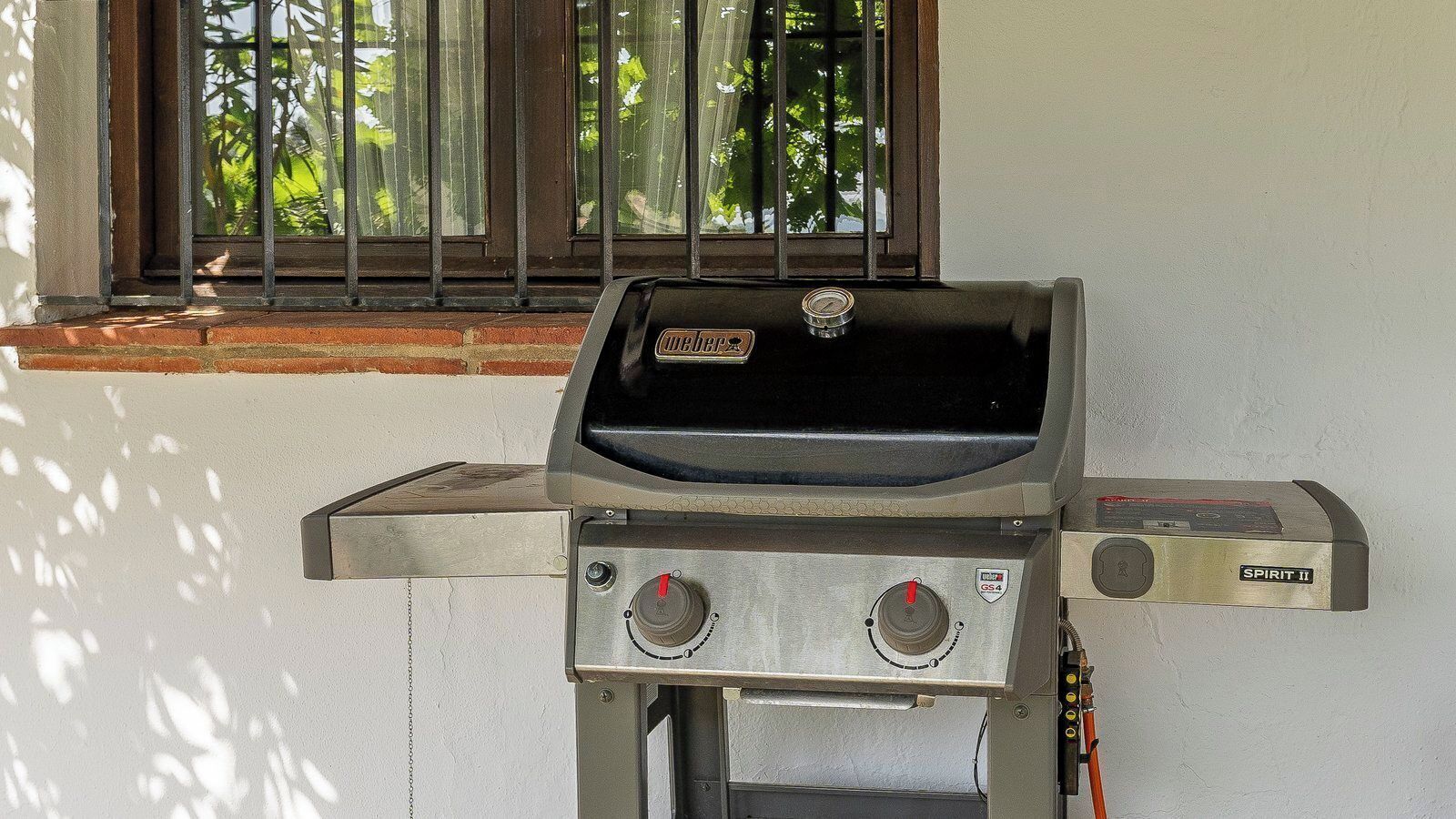 Una parrilla Weber Spirit II con dos perillas de control y una pantalla digital, ubicada frente a una pared blanca con una ventana al fondo.