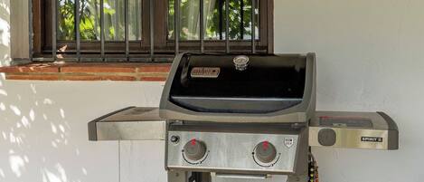 Una parrilla Weber Spirit II con dos perillas de control y una pantalla digital, ubicada frente a una pared blanca con una ventana al fondo.