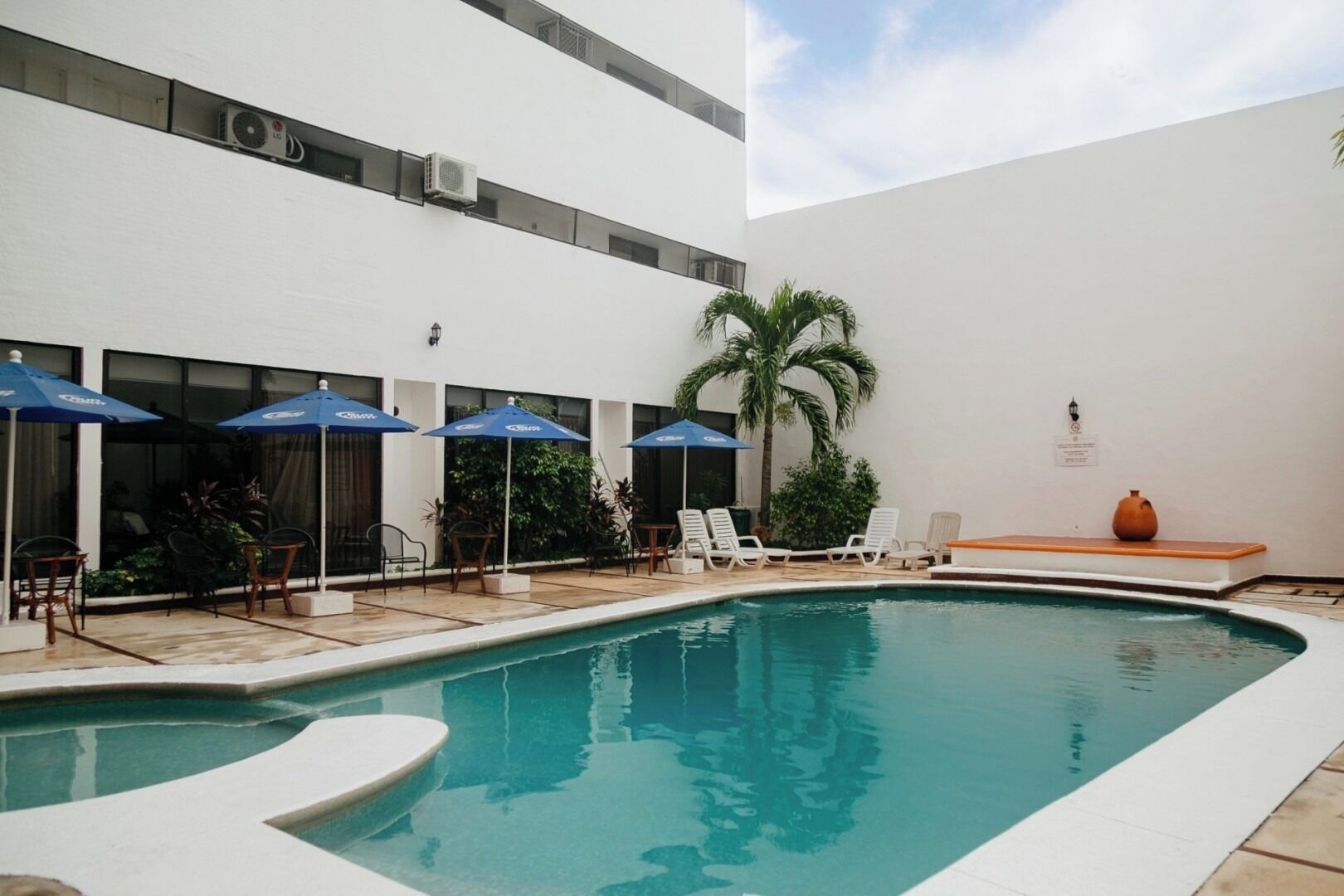 A swimming pool with lounge chairs and umbrellas, surrounded by a white building with air conditioning units.
