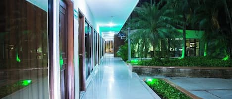 Corredor de hotel com portas de vidro, piso ladrilhado e palmeiras iluminadas por luz verde.