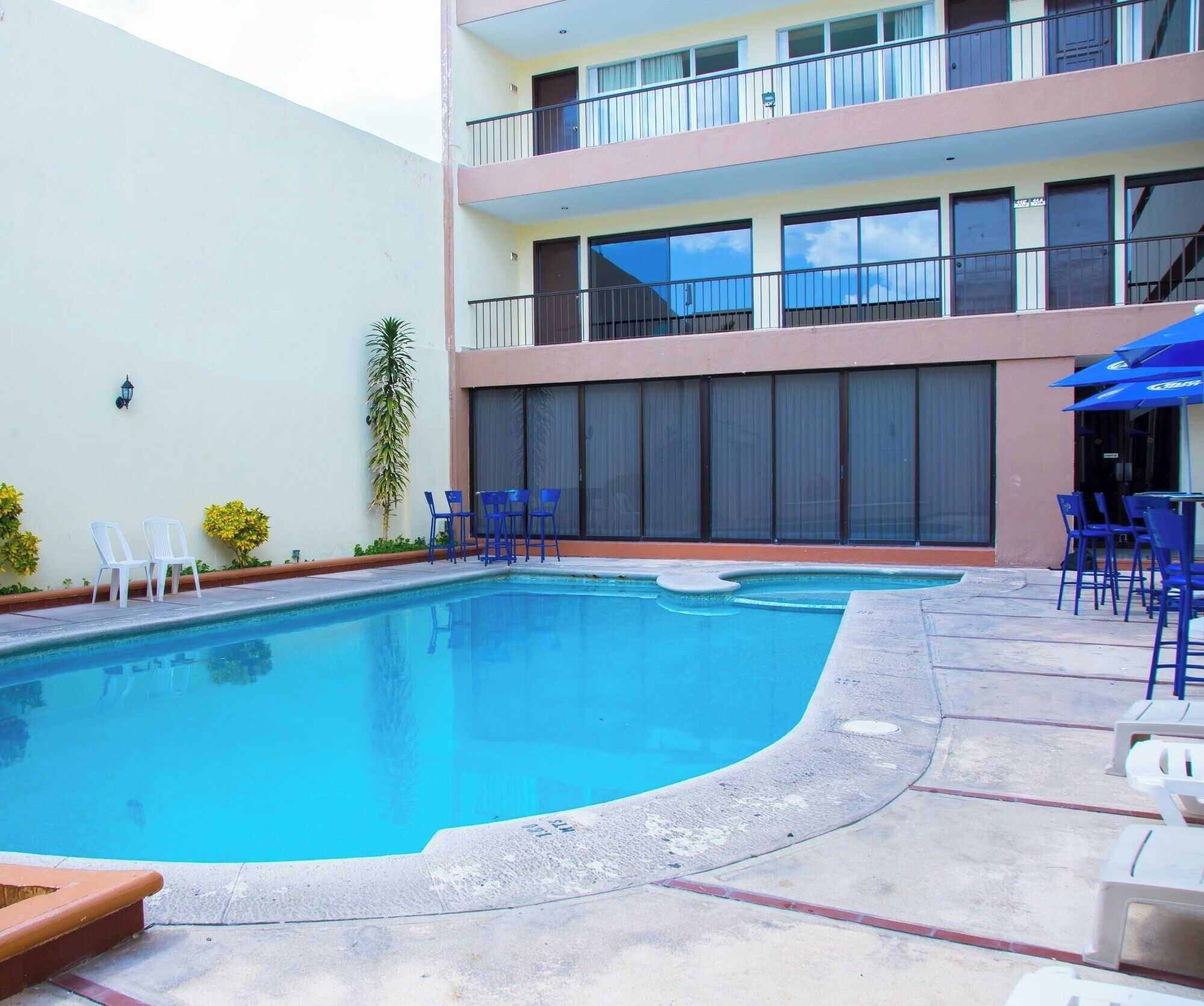 A swimming pool with a curved edge, surrounded by a patio area with chairs and tables. There is a building with balconies and large windows in the background.