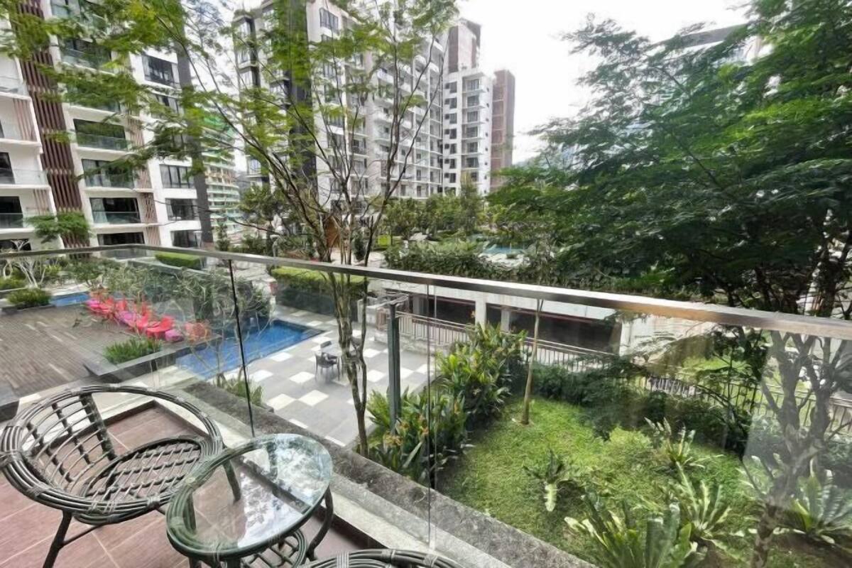 A balcony with glass railings, a small table, and chairs, overlooking a courtyard with plants and a pool.