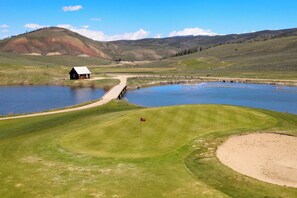 Un campo de golf con un estanque, un pequeño edificio y colinas onduladas al fondo.