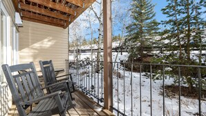 A wooden deck with two chairs and a table, a metal railing, and a view of a snowy landscape with trees. - Cozy Slope Slide Granby Condo (Granby)