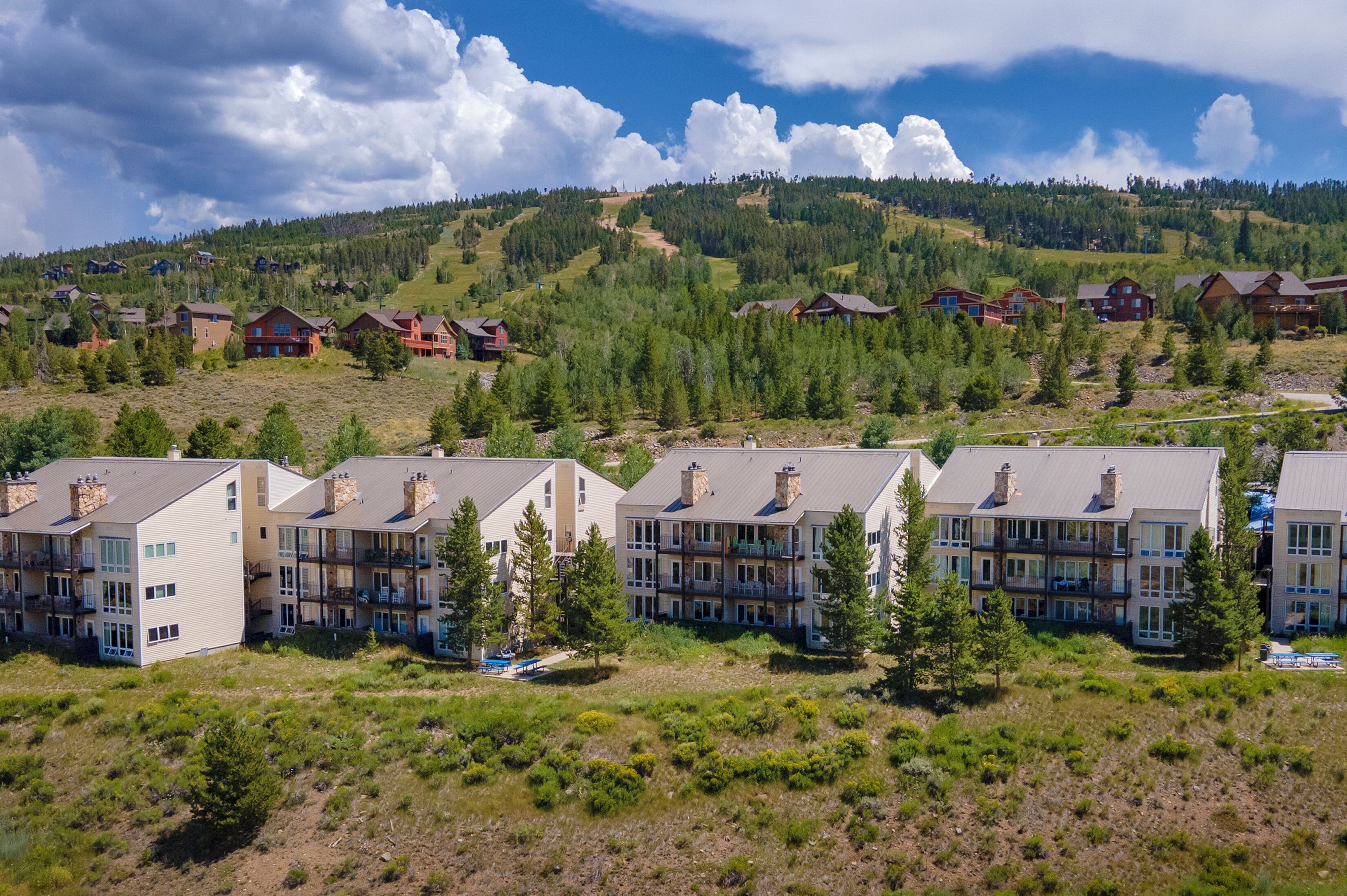 A cluster of multi-story buildings with balconies, set against a backdrop of a mountainous landscape with dense forests.
