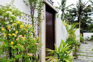 A white building with a dark wooden door, surrounded by lush greenery and a white lattice fence. - VIP Villas and Wellness Resort in Kuta Lombok (Pujut)