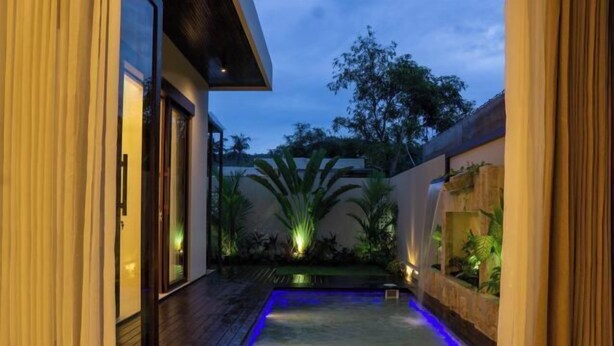 Un espace piscine extérieur moderne, doté d’une terrasse en bois, d’une petite cascade et d’un éclairage d’ambiance.