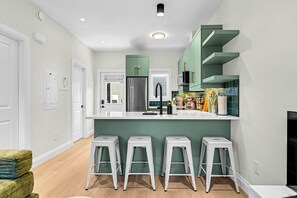Une cuisine moderne avec des meubles verts, un réfrigérateur en acier inoxydable et un plan de travail blanc. On y trouve quatre tabourets blancs et une petite table.