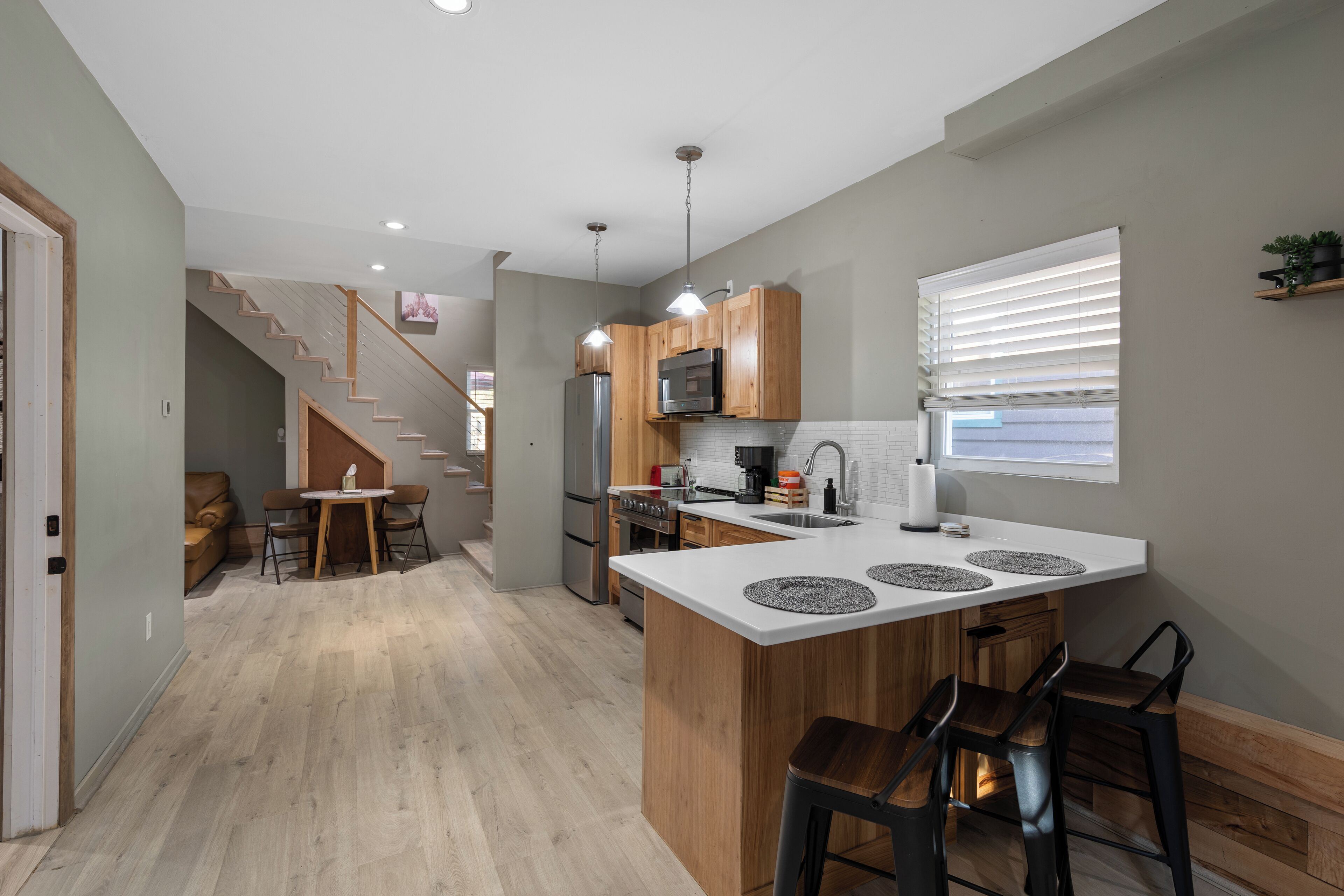 A modern kitchen with a central island, wooden cabinets, and a dining area with a staircase leading to another level.