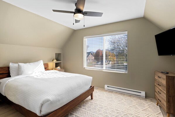A bedroom with a bed, a ceiling fan, a window with blinds, a TV mounted on the wall, and a wooden dresser. - Cozy Modern Home  (Lake Placid)