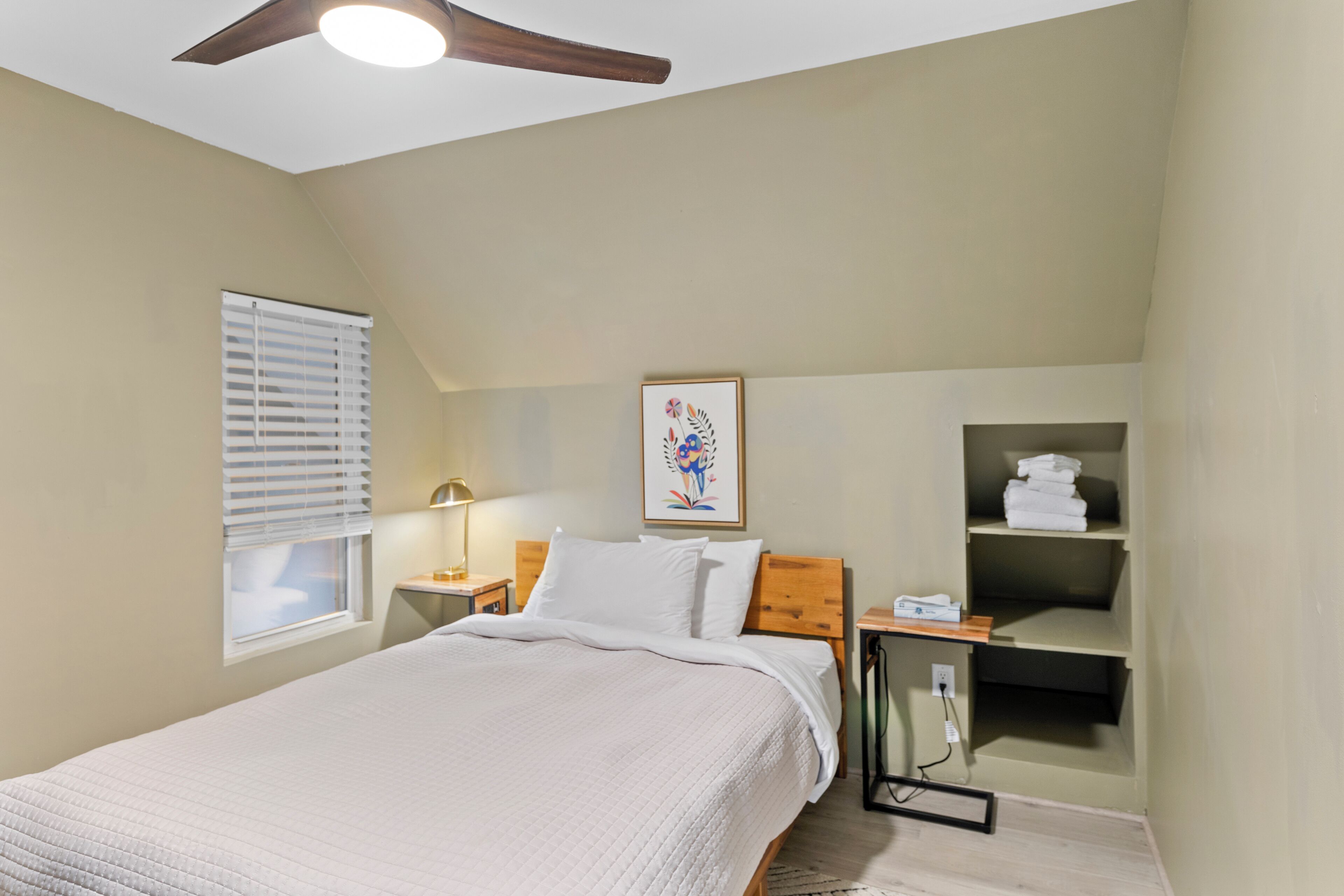 A bedroom with a bed, a nightstand, a wall-mounted shelf with towels, a ceiling fan, and a window with blinds.