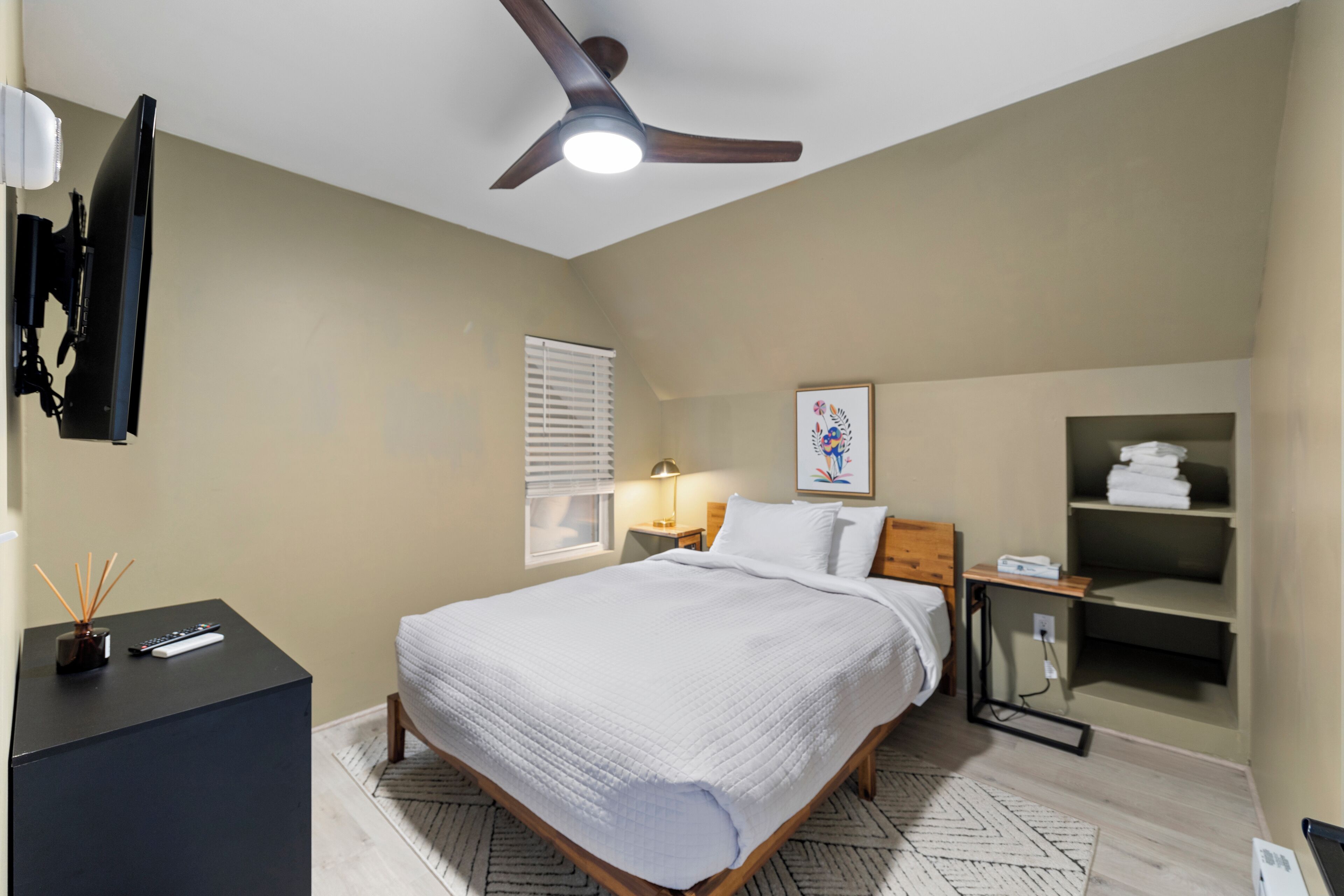 A bedroom with a bed, a ceiling fan, a bedside table with a lamp, a TV mounted on the wall, and a shelf with towels.