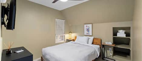 A bedroom with a bed, a ceiling fan, a bedside table with a lamp, a TV mounted on the wall, and a shelf with towels.