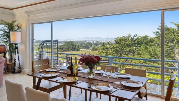 A dining area with a table set for four, large windows with a view of greenery and distant mountains, and a decorative vase with flowers. - Luxe Oceanview Game Room Retreat in Ideal Location (Daly City)