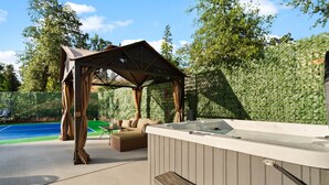 A hot tub with a wooden deck, a covered seating area, and a pool in the background.