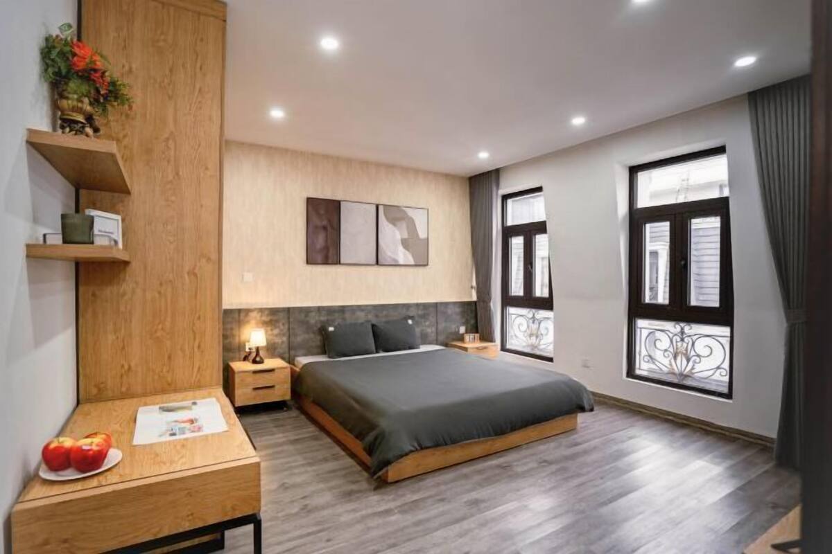 A modern bedroom with a large bed, wooden nightstands, a desk with apples and a lamp, and a window with a decorative iron grille.