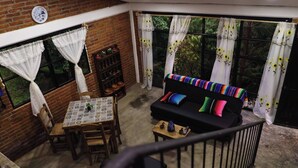 Una sala acogedora con un sofá negro, cojines de colores, una mesa de comedor con mantel estampado y una pared de ladrillo.