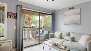 Una sala de estar con sofá, una mesa de centro y un ventilador de techo. Hay una puerta corrediza de vidrio que da a un área exterior con patio y vistas a la vegetación.