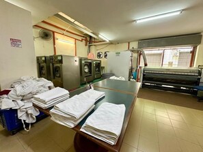 Une buanderie équipée de machines à laver et de séchoirs industriels, une grande table recouverte de serviettes pliées et une fenêtre avec des stores.