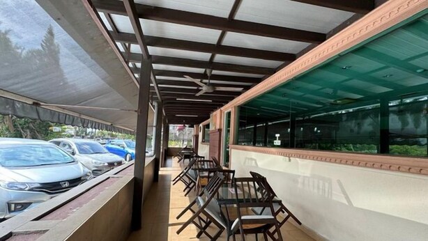 Un'area salotto esterna coperta con tavoli e sedie, una fila di auto parcheggiate e un edificio con ampie vetrate.
